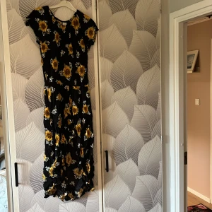 Svart långklänning med solrosor - Supersöt svart långklänning från Lindex med stora gula solrosor och små vita blommor. Klänningen har korta ärmar och ett flowy fall som ger en chill vibe. Perfekt för dig som gillar blommigt.Materialet är mjukt och skönt. Det finns även inbyggda shorts