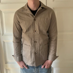 Beige overshirt från John Henric - Snygg beige overshirt från John Henric med klassisk krage och tre fickor framtill. Jackan har knäppning med knappar och är tillverkad i bomull. Perfekt för lager-på-lager och passar till jeans eller chinos. Jag är 183cm lång.