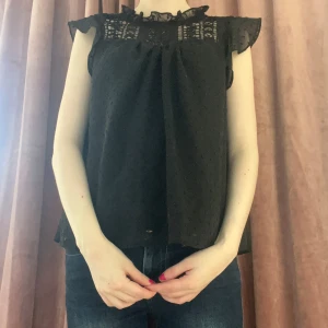 Svart blus med spets och volang - Supersöt svart blus med transparenta partier i spets upptill och volang vid halsen. Ärmlös modell med små prickar i tyget och lätt rynk vid axlarna. Perfekt att styla med jeans eller kjol för en trendig look.