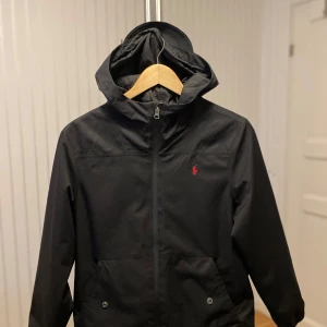 Svart vindjacka från Polo Ralph Lauren - Svart vindjacka från Polo Ralph Lauren med huva och dragkedja framtill. Jackan har en röd broderad logga på bröstet, två fickor med knapp och elastiska muddar vid ärmsluten. Perfekt för blåsiga dagar och enkel att matcha med allt.