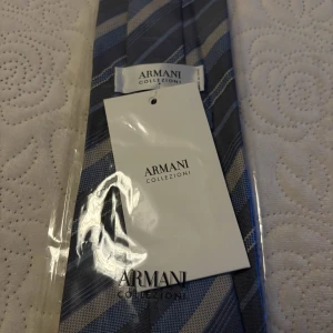 Armani Collezioni – ny blå slips i 100% siden. - Stilren och lyxig slips från Armani Collezioni, med vackert mönster i blå och grå toner. En klassisk accessoar som passar lika bra till kontoret som till festliga tillfällen.  Specifikationer:  Märke: Armani Collezioni  Färg: Blå/grå med diagonalrandigt mönster  Material: 100% siden  Skick: Ny och oanvänd  Förpackning: Originalförpackning med etikett kvar  Tillverkad i Italien  Högkvalitativ slips med sofistikerad design – perfekt present eller till dig själv!