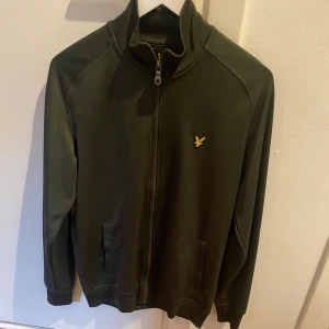 Mörkgrön ziptröja från Lyle & Scott - Snygg mörkgrön ziptröja från Lyle & Scott med gul logga på bröstet. Tröjan har hög krage, hel dragkedja och två fickor framtill. Perfekt för dig som gillar en clean och sportig stil.