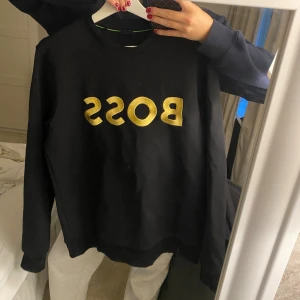 Svart BOSS sweatshirt med guldtryck - Svart sweatshirt från BOSS med stort, glansigt guldtryck på bröstet. Klassisk rund halsringning och långa ärmar. Tillverkad i mjuk bomull som är skön att ha på sig. Perfekt för dig som vill sticka ut med en clean och lyxig look.