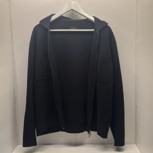 Svart overshirt från Massimo Dutti M - Svart overshirt från Massimo Dutti i storlek M. Tröjan har dragkedja framtill, två stora fickor på bröstet och huva. Materialet är mjukt och känns lite stickat, perfekt för kyliga dagar. Enkel och stilren design som funkar till det mesta.