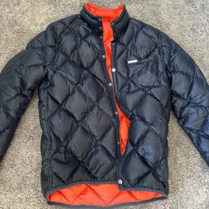 Quiltad dunjacka från Peak Performance - Snygg quiltad dunjacka från Peak Performance i mörkblått med orange foder. Jackan har hög krage, tryckknappar och en stilren look. Perfekt för kyliga dagar och enkel att matcha med olika outfits.