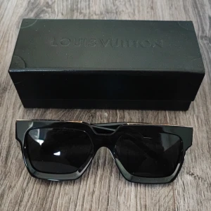 Svarta solglasögon - Säljer ett par svarta solglasögon, modell Pont Neuf. De har en fyrkantig form med breda bågar och detaljer i guld längs ovansidan och på sidorna.. Glasögonen är tillverkade i blankt syntetmaterial och har mörka linser.