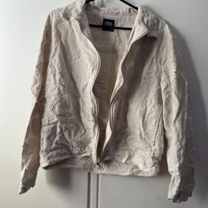 Beige strukturerad jacka från Zara - Säljer en beige jacka från Zara i storlek S. Jackan har en unik strukturerad yta med råa detaljer och dragkedja framtill. Den har klassisk krage och långa ärmar, perfekt för dig som gillar en lite annorlunda look.