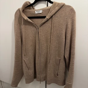 Beige kashmir hoodie från Rovoír - Mysig beige hoodie från Rovoír i kashmir material. Tröjan har huva, dragkedja framtill och två fickor. Perfekt för lager-på-lager och chill dagar. Långärmad och med snygg, enkel design som funkar till det mesta.