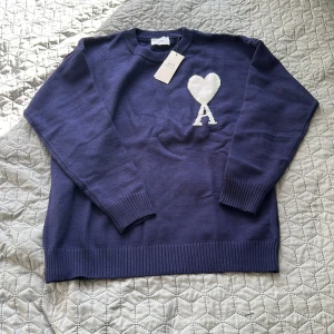 Marinblå stickad tröja från Ami - Snygg marinblå stickad tröja från Ami med ett stort vitt hjärta och bokstaven A på bröstet. Tröjan har rund halsringning, ribbade muddar och är långärmad. Perfekt för dig som vill ha en stilren men ändå unik look.