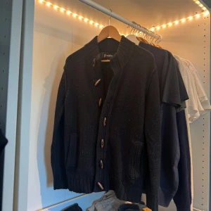 Svart stickad kofta från 7camicie - Svart stickad kofta från 7camicie med grov ribbad krage och snygga träknappar. Koftan har två fickor framtill och en liten patch på ärmen. Perfekt för lager-på-lager och chill höst- eller vinterstil.