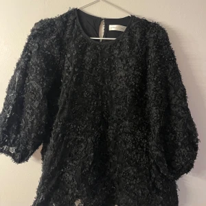 Svart blus - Supersnygg svart blus med 3D-blommor över hela tyget. Blusen har rund halsringning och trekvartsärmar, perfekt för att sticka ut med en unik textur. Materialet känns lyxigt och ger en cool look till outfiten.