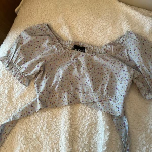 Blommig omlottopp  - Supersöt ljusblå omlottopp med små blommor i lila, rosa och gult. Toppen har puffiga korta ärmar, resår vid axlar och midja samt knytband i midjan som ger en snygg rosett bak. Perfekt för en somrig look!