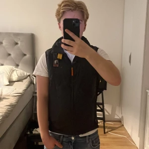 Svart dunväst Parajumpers XS - Säljer en svart dunväst från Parajumpers i storlek XS. Västen har huva, dragkedja framtill, klassisk Parajumpers-logga på bröstet och en gul detalj vid axeln. Perfekt att slänga över en hoodie eller t-shirt för en cool look. Västen har ett litet hål i fickan som inte märks. Passar 165-173