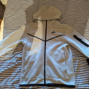 Grå Nike hoodie med dragkedja - Säljer en grå hoodie från Nike med hel dragkedja och svart logga på bröstet. Tröjan har huva, långa ärmar och en praktisk ficka med dragkedja på ena ärmen. Perfekt för chill eller träning.
