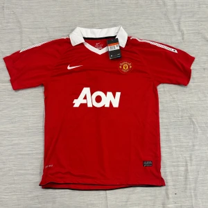 Manchester United röd Nike fotbollströja L från säsongen 10/11 - Manchester Uniteds officiella matchtröja från Nike i röd Dri-FIT polyester med vit krage och vita detaljer. Klassisk klubbmärke på bröstet, stor AON-logga och Nike Swoosh. Perfekt för dig som vill representera United på läktaren eller planen.