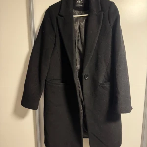 Svart kappa från Zara - Stilren svart kappa från Zara med klassisk krage och enkel knäppning framtill. Jackan har två fickor och är helfodrad, perfekt för dig som gillar en clean och tidlös look. Passar till allt och ger en snygg siluett.