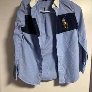 Blå skjorta från Ralph Lauren M - Snygg blå skjorta från Ralph Lauren med vit krage och mörkblå panel över bröstet. Ikonisk broderad polospelare på vänster sida. Skjortan har långa ärmar och knäppning framtill. Perfekt för dig som gillar klassisk men sportig stil.
