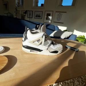 Nike Air Jordan 4 Retro vit/svart - Nike Air Jordan 4 Retro i vitt och svart med klassisk meshpanel, detaljer i mocka och skinn samt ikoniska Jordan-loggan på plösen. Snygg kontrast mellan svart och vitt, med coola plastdetaljer och synlig Air-sula. Perfekta för dig som gillar streetwear och sneakers med attityd.
