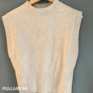 Beige stickad väst Pull&Bear XS - Beige stickad väst från Pull&Bear i storlek XS. Modellen är ärmlös med breda ribbade kanter vid ärmhål, halsringning och nederkant. Perfekt att slänga över en skjorta eller t-shirt för en chill och trendig look.
