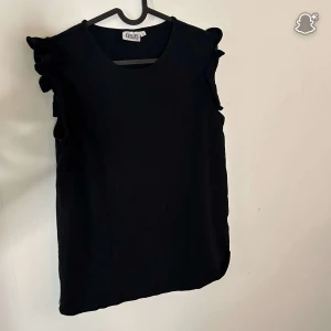 Svart ärmlös topp med volang från Dea - Säljer en svart ärmlös topp från Dea i storlek L. Toppen har volangdetaljer på axlarna som ger en extra touch och en rund halsringning. Perfekt att styla med jeans eller kjol för en snygg look.