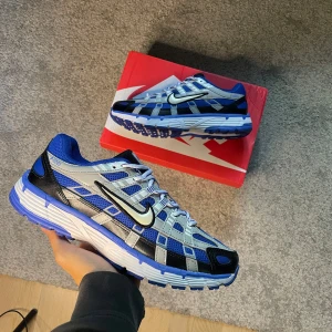 Nike P-6000 racer blue flat silver  - Nike P-6000 sneakers i blått, silver och svart med mesh och syntetpaneler. Skorna har vit och blå sula, svart Swoosh och klassisk snörning. Perfekt för dig som gillar retrovibbar och sportig stil. Snygga detaljer och bekväm passform.