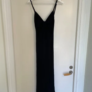 Svart långklänning med rosettdetalj - Säljer en svart långklänning med smala axelband och snygg rosettdetalj framtill. Klänningen har en figurnära passform och är ärmlös, perfekt för dig som gillar stilrena och trendiga plagg. Materialet känns mjukt och stretchigt.