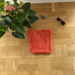 Röd t-shirt från Polo Ralph Lauren - Snygg röd t-shirt från Polo Ralph Lauren med klassisk grön logga broderad på bröstet. T-shirten har rund hals och korta ärmar, perfekt för chill dagar. Tillverkad i mjuk bomull som känns skön mot huden. Enkel och stilren design som funkar till jeans eller shorts.