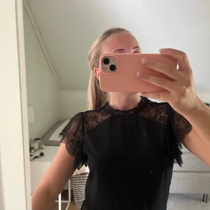 Svart blus med spetsdetaljer - Supersnygg svart blus med transparent spets upptill och på ärmarna. Blusen har korta, volangiga ärmar och en hög hals med spetskant. Knappt använd.
