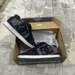 Svarta DC sneakers med grå detaljer - Snygga svarta DC sneakers med stora grå loggor på sidorna och vit sula. Skorna har snörning och en klassisk, chunky siluett som passar perfekt till streetwear. Materialmix av mocka och textil ger en cool kontrast. Kommer i originalkartong.