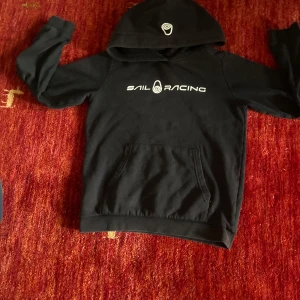 Svart hoodie från Sail Racing - Svart hoodie från Sail Racing med vit logga på bröstet och huvan. Klassisk känguruficka framtill och mjukt material som passar perfekt till chill dagar. Snygg och enkel design som funkar året runt.