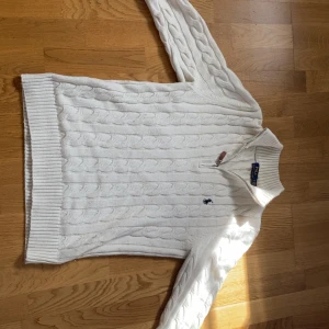 Vit kabelstickad tröja Polo Ralph Lauren - Vit kabelstickad tröja från Polo Ralph Lauren med halv dragkedja och klassisk broderad logga på bröstet. Tröjan har ribbade muddar och hög krage, perfekt för lager-på-lager. Snygg och tidlös design som funkar året om.