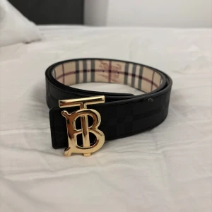 Svart Burberry bälte med guldspänne - Snyggt svart bälte från Burberry med klassiskt rutmönster och ett stort, glänsande guldspänne med TB-logga. Insidan har den ikoniska Burberry-rutan i beige, rött och svart. Tillverkat i exklusivt skinn och perfekt för att lyfta vilken outfit som helst.