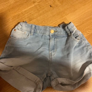 Ljusblå jeansshorts från Lindex - Snygga ljusblå jeansshorts från Lindex med klassisk femficksdesign och uppvikta benslut. Shortsen har guldiga detaljer vid knapparna och en bekväm midja med resår baktill. Perfekta för chill dagar och sommarhäng. JUSTERBAR VID MIDJAN!