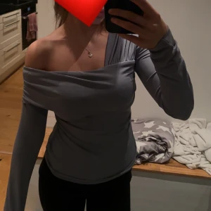 Grå oneshoulder topp med lång ärm - Säljer en stilren grå topp med oneshoulder-design och lång ärm. Toppen har en snygg drapering över axeln och sitter tight över kroppen. Perfekt för dig som vill ha en unik och trendig look.