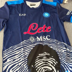 Napoli Maradona 10 EA7 fotbollströja - Snygg Napoli fotbollströja från EA7 med Maradona och nummer 10 på ryggen. Tröjan är marinblå med ljusblå detaljer, vita tryck och coolt grafiskt mönster framtill. Kortärmad modell i lätt och ventilerande material, perfekt för match eller träning.