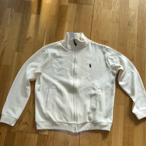 Vit ziptröja från Ralph Lauren - Vit långärmad ziptröja från Ralph Lauren med hög krage och klassisk broderad logga på bröstet. Tröjan har två fickor framtill och är tillverkad i mjuk bomull. Perfekt för en clean och stilren look. org1nal helt ny