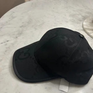 Svart keps från Gucci med logomönster - Snygg svart keps från Gucci med diskret GG-logomönster över hela kepsen. Klassisk böjd skärm och justerbar baksida. Materialet känns slitstarkt och har en lyxig känsla, perfekt för dig som gillar designeraccessoarer.