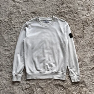 Stone Island sweatshirt - Vit crewneck sweatshirt från Stone Island med klassisk rund hals och ribbade muddar. Tröjan har den ikoniska svarta patchen med gul broderad kompass på vänster ärm. Tillverkad i mjuk bomull, perfekt för chill dagar och streetwear-stil.