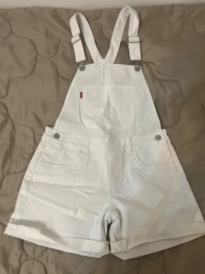 Vita Levi's shortalls i jeans - Säljer ett par vita shortalls från Levi's i jeansmaterial. De har justerbara hängslen, bröstficka framtill och klassiska femficksdetaljer. Shortsen har uppvikta benslut och Levi's patch bak. Perfekta för en avslappnad och cool stil.