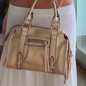 Beige handväska med dragkedja och detaljer - Snygg beige handväska i skinnimitation med både handtag och axelrem. Väskan har coola spänndetaljer, dragkedja framtill och dekorativa tofsar. Perfekt storlek för att få plats med allt du behöver och passar till många olika outfits.