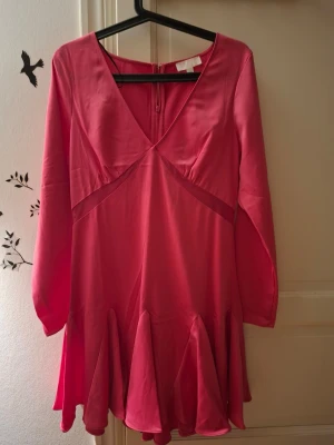 Rosa kortklänning med volang - Supersöt rosa kortklänning med v-ringning och långa ärmar. Klänningen har volangdetaljer nertill och är tillverkad i ett glansigt, mjukt material som ger en lyxig känsla. Perfekt för dig som vill sticka ut med en färgstark look.