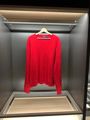 Röd v-ringad tröja Polo Ralph Lauren - Riktigt snygg röd v-ringad tröja från Polo Ralph Lauren i mjuk pima bomull. Klassisk logga broderad på bröstet och ribbade muddar vid ärmslut och nederkant. Perfekt för dig som vill ha en stilren och tidlös look.                                                Längd:69cm.     Bredd:48cm