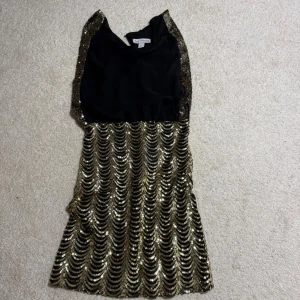 Svart och guld paljettklänning XS - Supersnygg kort klänning från Model Behaviour i svart med guldiga paljetter i vågmönster. Klänningen har ärmlös design och en riktigt festlig look som verkligen sticker ut. Perfekt för dig som vill glänsa på nästa event!