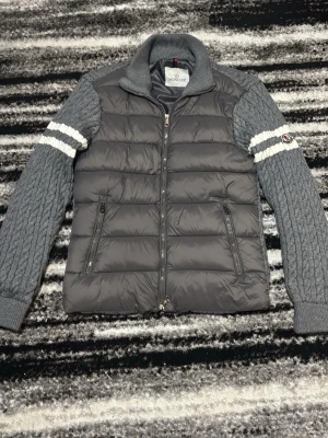 Grå pufferjacka från Moncler - Säljer en grå pufferjacka från Moncler med stickade ärmar och krage. Ärmarna har kabelstickat mönster och vita ränder med Moncler-logga på vänster arm. Jackan har dragkedja framtill och två fickor med dragkedja. Perfekt för dig som vill ha en unik och snygg jacka.