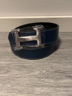 H-bälte  - Snyggt bälte i blått och svart skinn med ikoniskt,H-spänne i silverfärgad metall. Bältet har stilrena, kontrastsömmar och klassisk design som lyfter, vilken outfit som helst. Strl 110cm