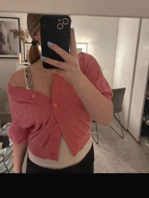 Rosa kofta med kort ärm och knappar - Söt rosa kofta med korta ärmar och dekorativa hålstickade detaljer. Koftan har knappar framtill och en bred ribbad kant. Perfekt att bära off-shoulder för en trendig look. Materialet känns mjukt och är stickat. 