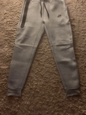 Grå mjukisbyxor från Nike - Säljer ett par grå mjukisbyxor från Nike med svart logga på vänster lår och snörning i midjan. Byxorna har en dragkedjeförsedd ficka på höger sida och muddar vid bensluten. Perfekta för chill eller träning.