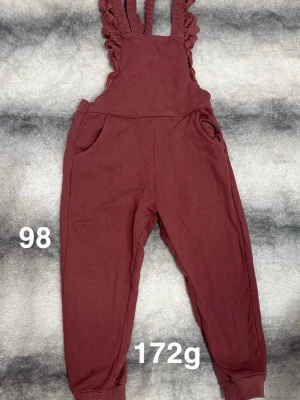 Vinröd byxdress med volangaxel - Supersöt vinröd byxdress med volangprydda axelband. Byxdressen har fickor på sidorna och muddar vid bensluten för en bekväm passform. Perfekt för dig som gillar en lekfull och trendig stil.🌸 Använd en gång. Storlek 98