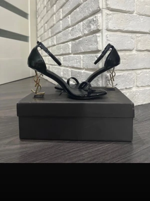 Yves Saint Laurent  klackskor - Eleganta svarta klackskor från Yves Saint Laurent (YSL) med ikonisk guldfärgad klack formad som YSL-loggan. Skorna har en stilren, lackad finish och smala remmar som ger ett exklusivt och sofistikerat intryck. Perfekta för fest, middag eller andra tillfällen där du vill sticka ut med en lyxig detalj. En tidlös designerklack som kombinerar mode och elegans.