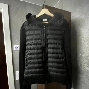 Moncler cardigan - Svart pufferjacka från Moncler med stickade ärmar och quiltad front. Jackan har huva, dragkedja framtill och två fickor med dragkedja. Moncler-logga på ena ärmen. Perfekt för kyliga dagar. Jackan är i väldigt bra skick utan defekter.